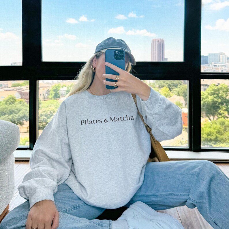 Puede incluir: Una sudadera gris claro con el texto "Pilates & Matcha" en negro. La persona lleva una gorra azul, vaqueros azules y zapatillas blancas. El fondo muestra una vista de la ciudad con cielo azul.