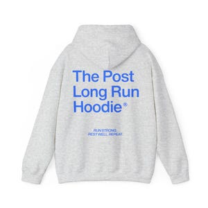 May include: A light gray hoodie with a hood and long sleeves. The back of the hoodie features the text "The Post Long Run Hoodie" in blue, with the words "Run Strong. Rest Well. Repeat." printed below.