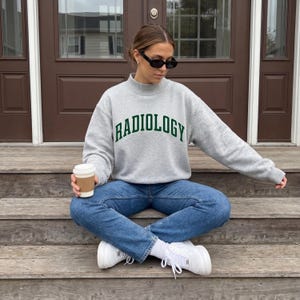 Peut inclure: Un sweat-shirt gris avec le mot "RADIOLOGY" en lettres vertes. Le sweat-shirt a un col montant et est porté avec un jean bleu et des baskets blanches. Une personne tient une tasse de café.