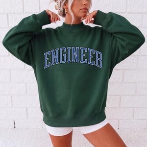 Peut inclure: Un sweat-shirt à manches longues vert foncé avec un col montant et des poignets côtelés. Le mot ENGINEER est imprimé sur la poitrine dans une police universitaire arquée et audacieuse, avec des lettres bleues soulignées de blanc.