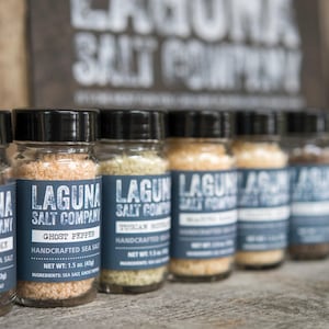 May include: A row of eight glass jars filled with different types of sea salt. Each jar has a black label with the text "Laguna Salt Company" and the name of the salt variety. The salts include "Humer Smoke", "Ghost Pepper", "Tuscan", "Black Truffle", "Maldon", "Fleur de Sel", "Sea Salt Flakes", and "Pink Himalayan".