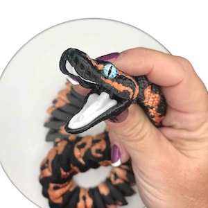 May include: A detailed, handmade snake-shaped accessory with black and orange patterns. The snake has a white mouth, blue eyes, and is held on a white plate. The accessory is likely a hair tie or scrunchie.