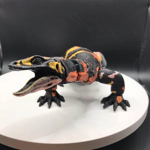 May include: A 3D-printed lizard model, predominantly black with orange and yellow patterns. The model has its mouth open, showing a pink tongue. It is displayed on a white circular platform, with a dark background.