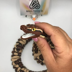 May include: A brown and tan snake-shaped ornament with a detailed head and open mouth. The snake has yellow eyes and is held in a hand. The background includes a business card with the text "Asterix Printing Co."