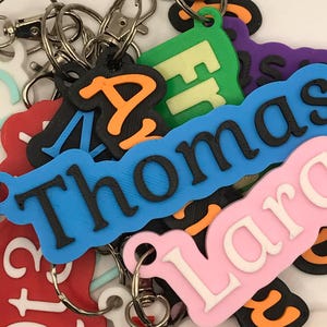 May include: A selection of personalised keyrings in various colours. The keyrings feature names and letters in black and orange, set against backgrounds of blue, pink, green, and red. Each keyring has a metal ring.