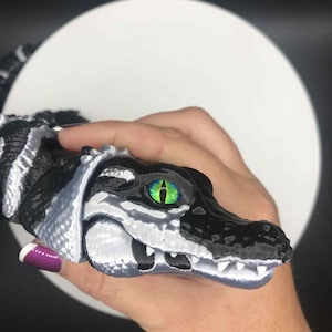 May include: A 3D-printed alligator figurine with a black and white body, a black head, and a green and yellow eye. The alligator's mouth is open, revealing small white teeth. The figurine is held in a hand.