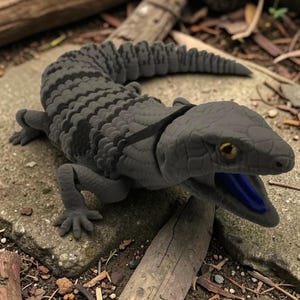 May include: A detailed, grey lizard sculpture with a blue tongue. The reptile has a textured body with dark grey stripes and is posed on a stone surface. The sculpture's eye is yellow.