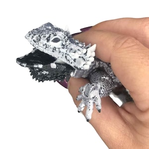 May include: A detailed, grey and white toy lizard with a textured, scaly appearance. The lizard has a black frill and is held in a hand against a white background. The toy has small, sharp spikes and claws.