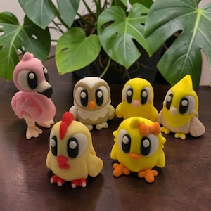 May include: A selection of handmade clay bird figurines, including a pink flamingo, an owl, and yellow chicks. The figurines have large eyes and are displayed on a dark brown surface, with a leafy green plant visible in the background.