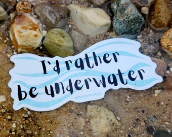 Könnte beinhalten: Ein weißer Aufkleber mit blauen Wellenlinien und dem Text "I'd rather be underwater" in Schwarz. Der Aufkleber befindet sich auf einem Bett aus kleinen, glatten, braunen Steinen.