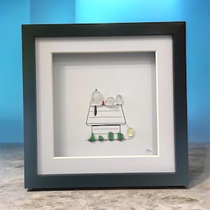Puede incluir: Obra de arte enmarcada que representa a Snoopy acostado en su caseta, con Woodstock cerca. La obra está creada con piedras lisas y redondeadas sobre un fondo blanco. El marco es de color gris oscuro.