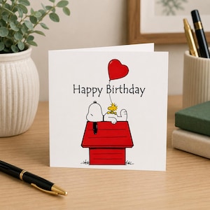 May include: A white square greeting card featuring a black and white illustration of Snoopy lying on a red doghouse with Woodstock. A red heart-shaped balloon is tied to the doghouse. The text Happy Birthday is printed in black above the characters.