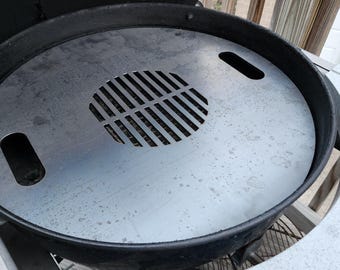 May include: A round, silver-colored grill grate with a central slotted vent and two handle cutouts. The grate sits inside a black grill, with a partially visible wooden deck in the background. The grill is designed for outdoor cooking.