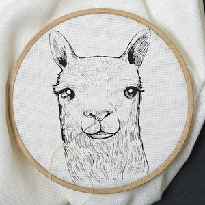 May include: A black and white line drawing of a llama's face is embroidered on a white fabric stretched in a wooden embroidery hoop. The hoop is on a black surface with gold embroidery thread, a spool of thread, and gold thimbles.