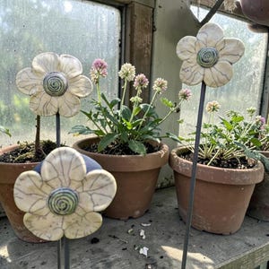 Könnte beinhalten: Cremefarbene Gartenstecker in Form von Blumen aus Keramik mit Craquelé-Finish und grünen Zentren. Die Stecker werden in Terrakotta-Töpfen mit blühenden Pflanzen ausgestellt und schaffen eine charmante Gartenszene.