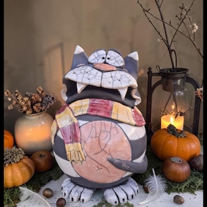 Könnte beinhalten: Eine Keramik-Katzenfigur mit rundem Körper, die ein dunkelgrau-weißes Streifenmuster, einen pfirsichfarbenen Bauch und einen gelb-rot gestreiften Schal aufweist. Die Katze hat ein breites, zahniges Grinsen, große Augen und eine Rissglasur.