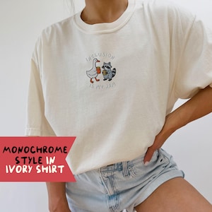 K&ouml;nnte beinhalten: Elfenbeinfarbenes T-Shirt mit einem gestickten Design einer Gans und eines Waschb&auml;ren, die Kaffeetassen halten, mit dem Text "Inclusion is my jam". Das Shirt hat kurze &Auml;rmel und einen Rundhalsausschnitt. Unten links im Bild steht der Text "Monochrome Style in Ivory Shirt".
