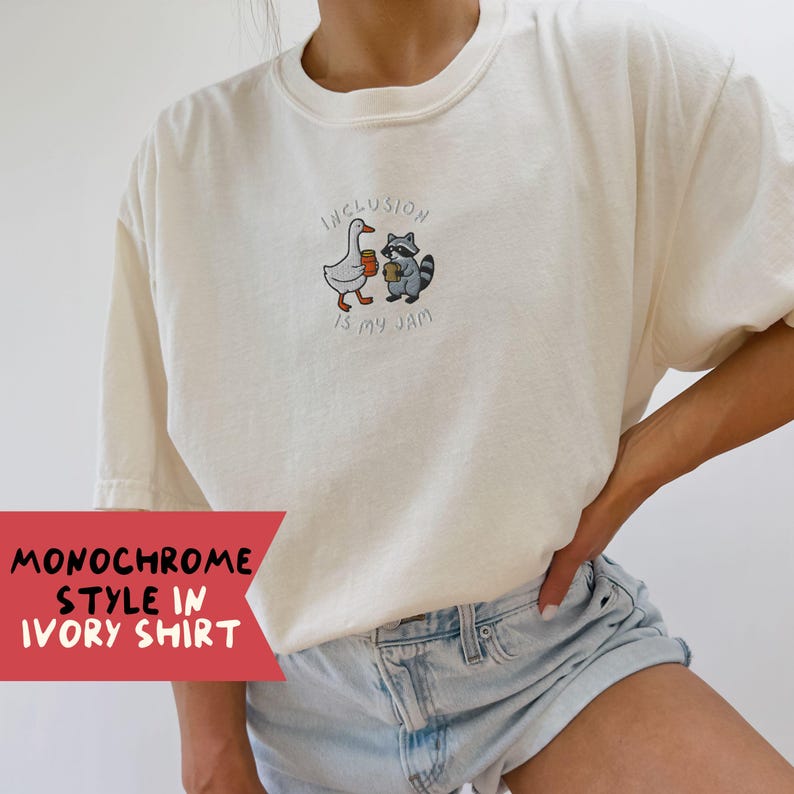 K&ouml;nnte beinhalten: Elfenbeinfarbenes T-Shirt mit einem gestickten Design einer Gans und eines Waschb&auml;ren, die Kaffeetassen halten, mit dem Text "Inclusion is my jam". Das Shirt hat kurze &Auml;rmel und einen Rundhalsausschnitt. Unten links im Bild steht der Text "Monochrome Style in Ivory Shirt".