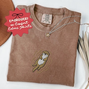 May include: A brown Comfort Colors t-shirt with an embroidered otter design. The shirt has the text "EMBROIDERY on Comfort Colors Shirts" on a red starburst. A silver necklace with a small pendant is on the shirt.