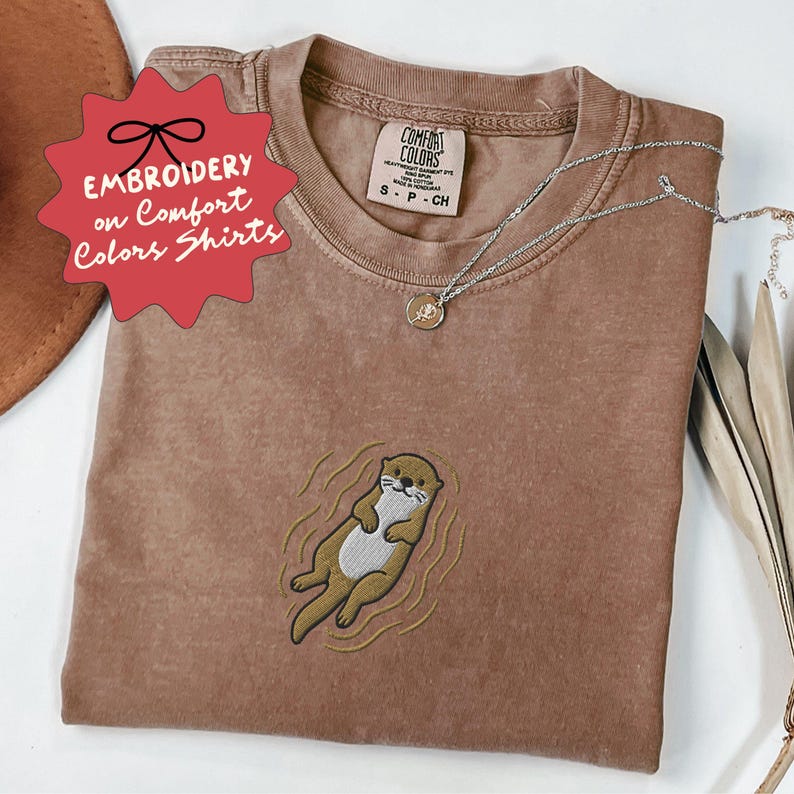 May include: A brown Comfort Colors t-shirt with an embroidered otter design. The shirt has the text "EMBROIDERY on Comfort Colors Shirts" on a red starburst. A silver necklace with a small pendant is on the shirt.