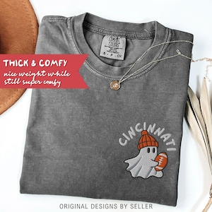 May include: A grey Comfort Colors t-shirt featuring an embroidered ghost wearing an orange beanie and holding a football, with the word "Cincinnati" above. The shirt has the text "Thick & Comfy" and "Original Designs by Seller".
