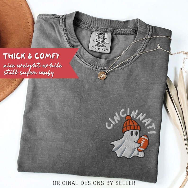 May include: A grey Comfort Colors t-shirt featuring an embroidered ghost wearing an orange beanie and holding a football, with the word "Cincinnati" above. The shirt has the text "Thick & Comfy" and "Original Designs by Seller".