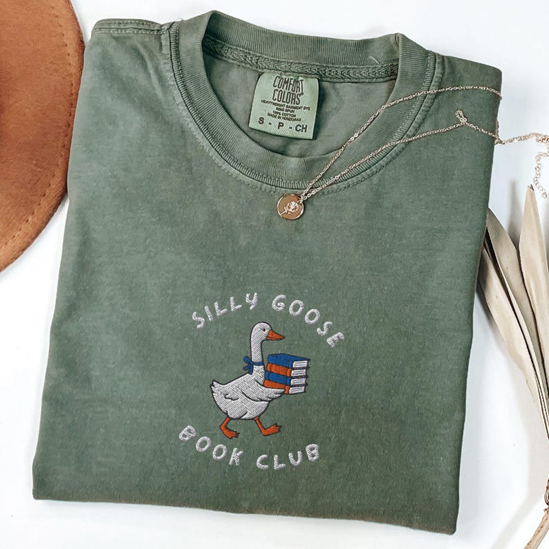 Może przedstawiać: Zielony T-shirt z haftem "Silly Goose Book Club" na przodzie. Projekt przedstawia kresk&oacute;wkową gęś niosącą stos książek.