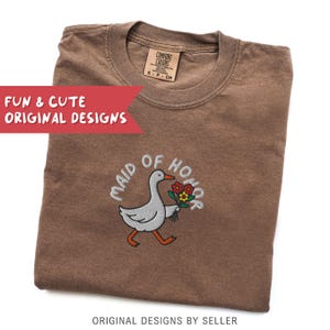 May include: A brown Comfort Colors t-shirt with the embroidered phrase "MAID OF HONOR" and a cartoon goose carrying flowers. The shirt also features the text "FUN & CUTE ORIGINAL DESIGNS" and "ORIGINAL DESIGNS BY SELLER".