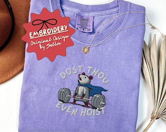 May include: A light purple t-shirt with an embroidered design of a possum lifting weights, with the text "DOST THOU EVEN HOIST". The shirt also has the text "EMBROIDERY Original Design by Seller" and "DIFFERENT COLORS AVAILABLE". A gold necklace is draped on the shirt.