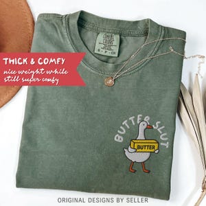 以下が含まれることがあります： オリーブグリーンのComfort Colors Tシャツ。刺繍された「BUTTER SLUT」の文字と、バターの塊を持った漫画のガチョウが描かれています。Tシャツには「THICK & COMFY」と「nice weight while still super comfy」の文字があります。金のネックレスがTシャツの上にかけられています。