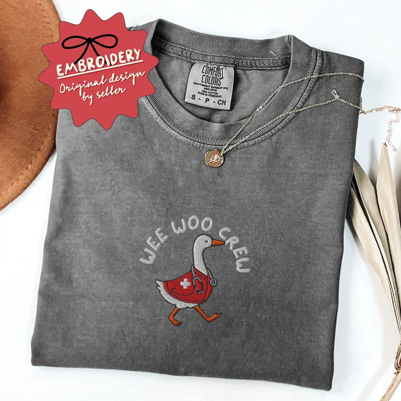 May include: A gray Comfort Colors t-shirt with the embroidered phrase "WEE WOO CREW" and a cartoon goose wearing a red medical coat. The shirt also has a small gold necklace and a red star-shaped tag that says "EMBROIDERY Original design by seller."