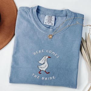 May include: A light blue t-shirt with a white embroidered goose wearing a veil and the text "Here Comes The Bride".