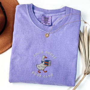 Może przedstawiać: Fioletowy T-shirt z białym haftem przedstawiającym gęś niosącą książki i tekst "Silly Goose Book Club".