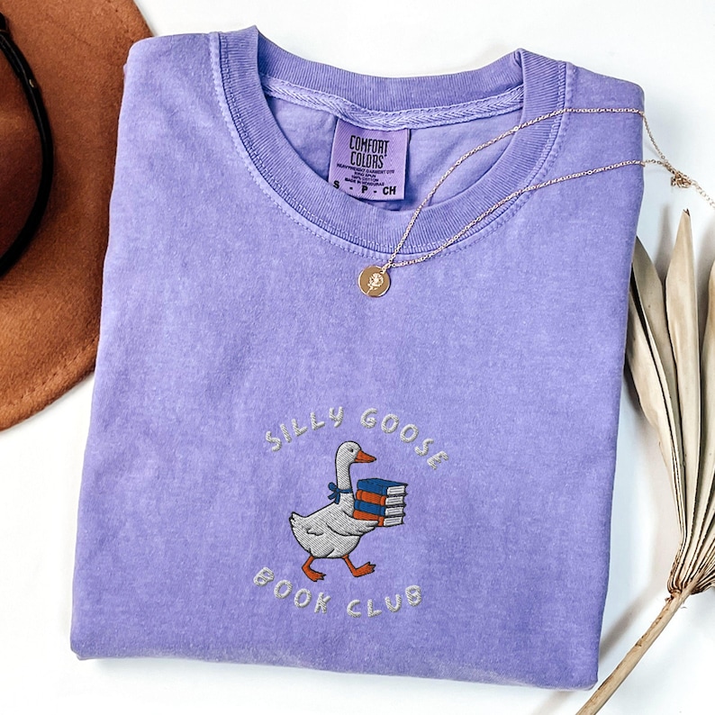 Może przedstawiać: Fioletowy T-shirt z białym haftem przedstawiającym gęś niosącą książki i tekst "Silly Goose Book Club".