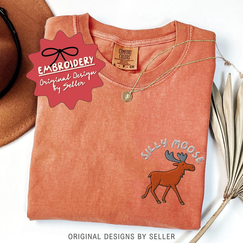 May include: A coral-colored t-shirt with the embroidered phrase "Silly Moose" and a cartoon moose design. A gold necklace and a red tag that says "Embroidery Original Design by Seller" are also visible.