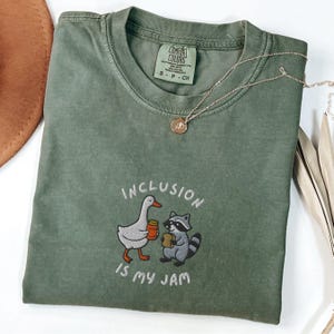 K&ouml;nnte beinhalten: Olivgr&uuml;nes T-Shirt mit dem gestickten Schriftzug "INCLUSION IS MY JAM". Das Design zeigt eine Cartoon-Gans und einen Waschb&auml;ren, die Marmeladengl&auml;ser halten. Eine goldene Halskette mit einem kleinen Anh&auml;nger liegt auf dem Shirt.