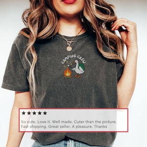 Op de afbeelding: Donkergrijs T-shirt met een geborduurd ontwerp met een gans met een rugzak die een marshmallow roostert boven een kampvuur. De tekst "CAMPING CREW" staat boven de gans. Het shirt is een comfortabele, casual stijl.