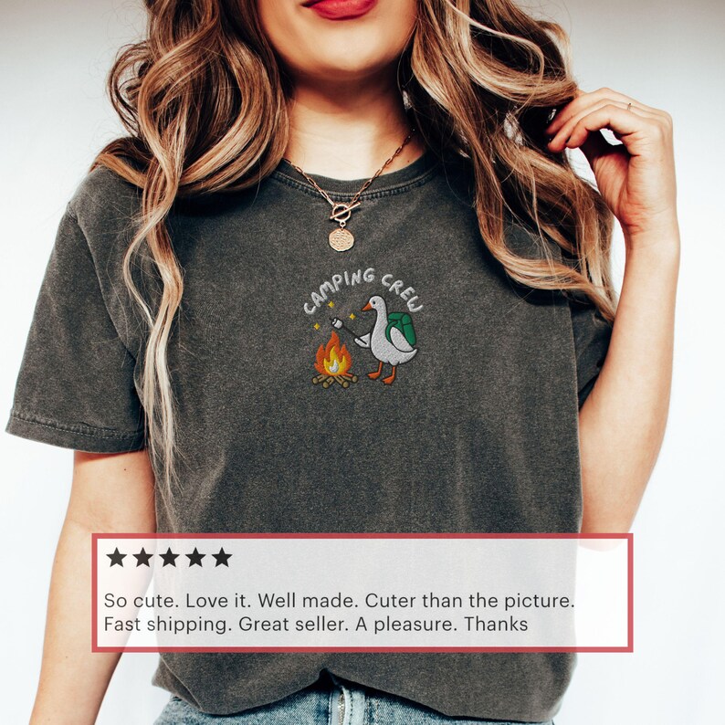 Op de afbeelding: Donkergrijs T-shirt met een geborduurd ontwerp met een gans met een rugzak die een marshmallow roostert boven een kampvuur. De tekst "CAMPING CREW" staat boven de gans. Het shirt is een comfortabele, casual stijl.