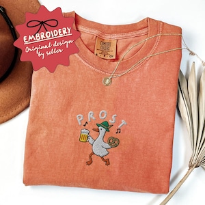 May include: Coral-colored t-shirt with an embroidered design featuring a cartoon goose wearing a green hat, holding a beer and a pretzel, with the word "PROST" above. A gold necklace and a red tag that says "EMBROIDERY Original design by seller" are also visible.