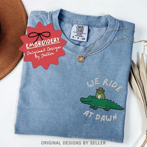 May include: A light blue t-shirt with an embroidered design featuring a green alligator and a frog wearing a hat, with the text "WE RIDE AT DAWN". The shirt also has a gold necklace and a red tag that says "EMBROIDERY Original Design by Seller".