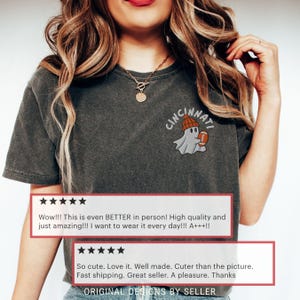 May include: Dark grey t-shirt featuring a Cincinnati ghost design. The ghost is wearing a beanie and holding a football. The shirt has a vintage appearance and includes customer reviews. The text "ORIGINAL DESIGNS BY SELLER" is at the bottom.
