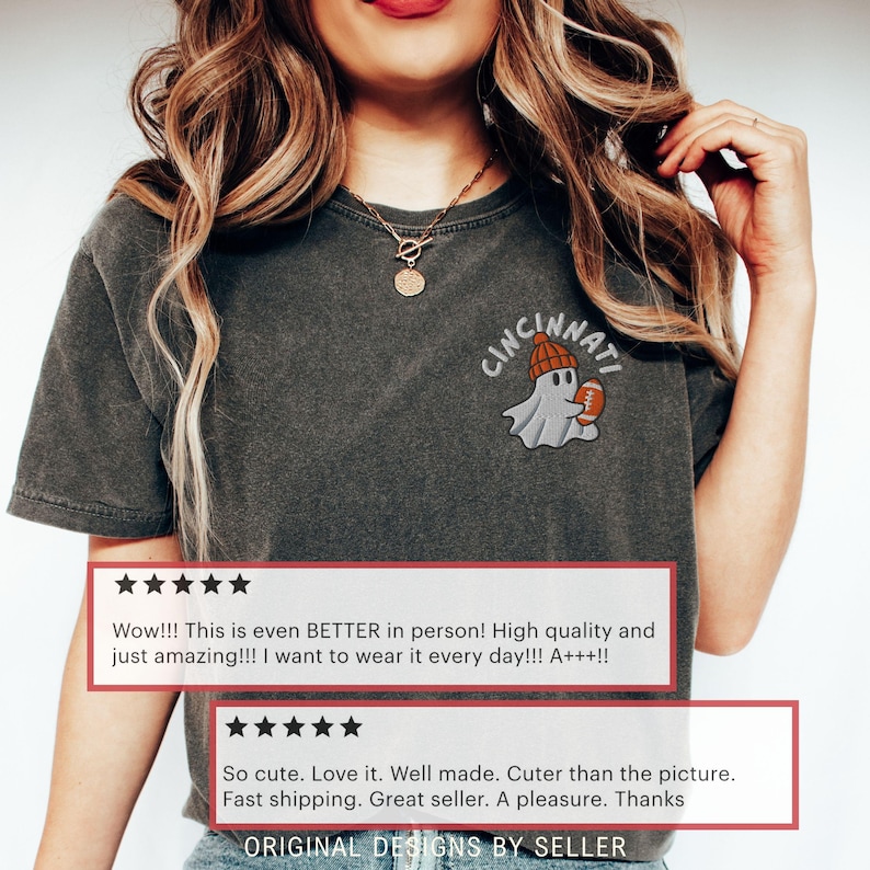 May include: Dark grey t-shirt featuring a Cincinnati ghost design. The ghost is wearing a beanie and holding a football. The shirt has a vintage appearance and includes customer reviews. The text "ORIGINAL DESIGNS BY SELLER" is at the bottom.