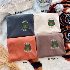 May include: Four folded t-shirts in pepper, ivory, yam, and espresso colors. Each shirt features an embroidered frog graphic with the text "WHY I OUGHTA". The shirts are displayed on a patterned surface.