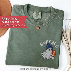 May include: Olive green t-shirt with a ghost wearing a blue and red beanie, holding a football, and the word "BUFFALO" embroidered above. The shirt also has the text "BEAUTIFUL FADED COLORS Effortlessly aesthetic". A gold necklace is draped across the shirt.