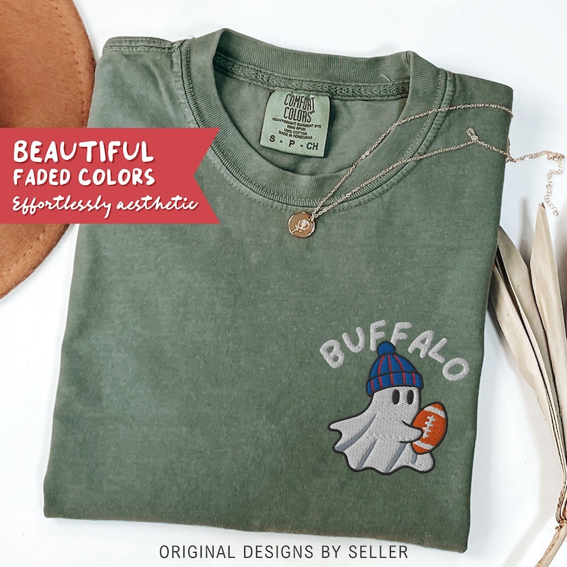 May include: Olive green t-shirt with a ghost wearing a blue and red beanie, holding a football, and the word "BUFFALO" embroidered above. The shirt also has the text "BEAUTIFUL FADED COLORS Effortlessly aesthetic". A gold necklace is draped across the shirt.