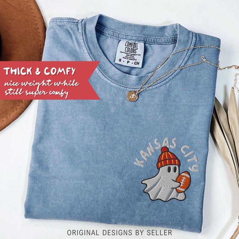 May include: A blue Comfort Colors t-shirt with a ghost wearing a red and orange beanie and holding a football. The text "Kansas City" is above the ghost. The shirt has the text "THICK & COMFY" and "ORIGINAL DESIGNS BY SELLER". A gold necklace is on the shirt.