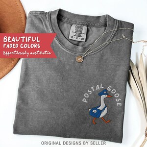 May include: A gray Comfort Colors t-shirt with the embroidered text "POSTAL GOOSE" and a cartoon goose wearing a postal worker hat and carrying a mail bag. The shirt also has the text "ORIGINAL DESIGNS BY SELLER".