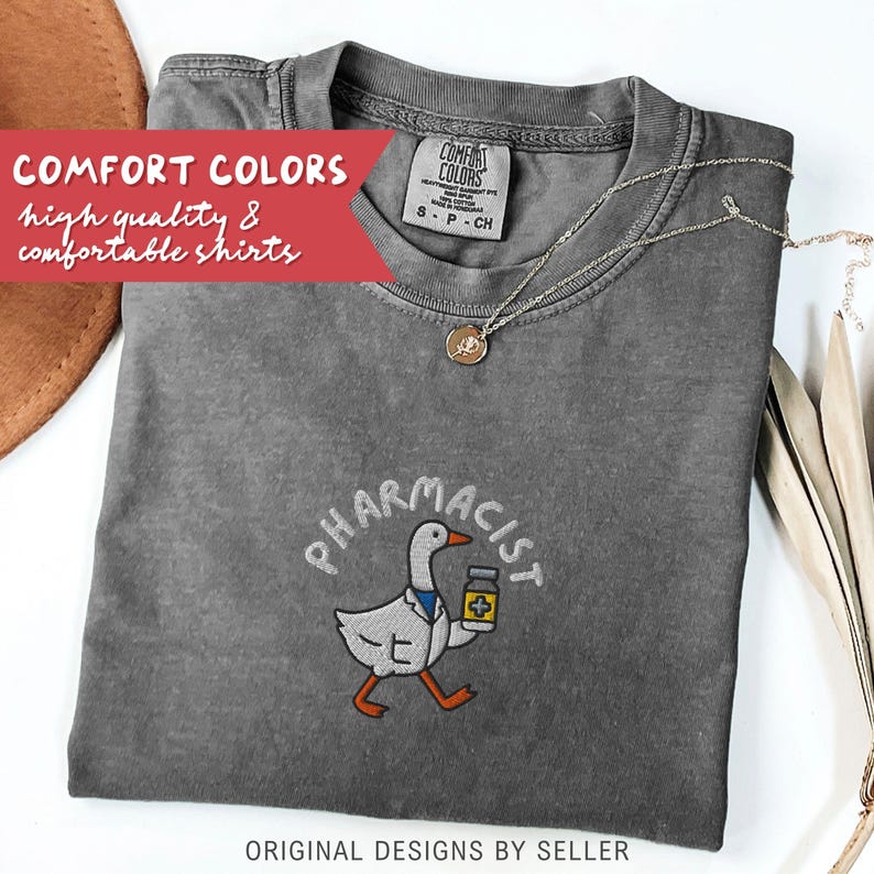 May include: A gray t-shirt with the word "PHARMACIST" embroidered above a cartoon goose holding a medicine bottle. The shirt has the text "COMFORT COLORS high quality & comfortable shirts" in a red banner. A silver necklace with a pendant is also visible.