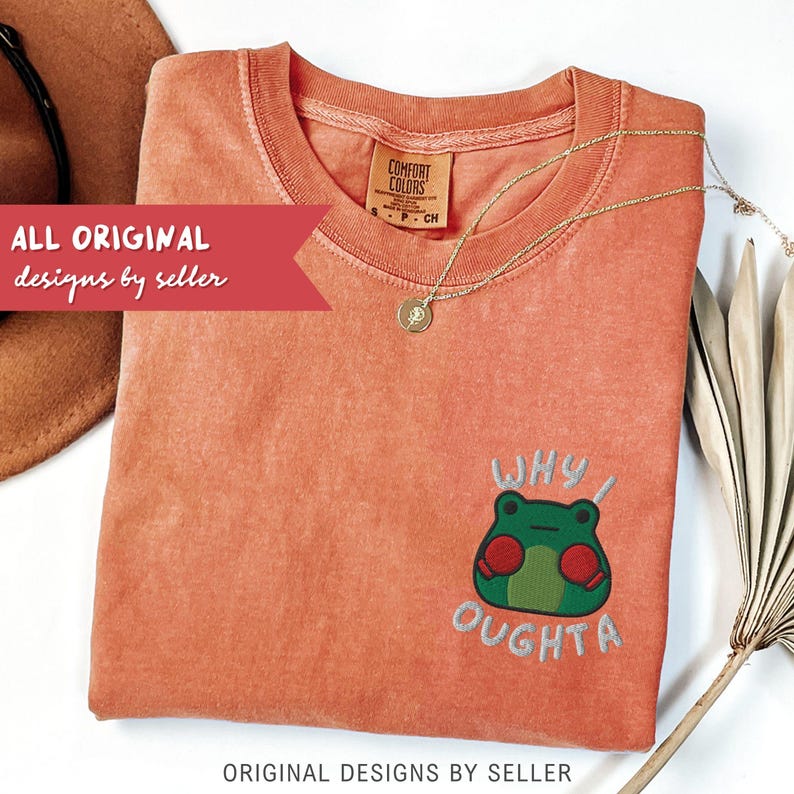 May include: Coral-colored t-shirt with a frog graphic and the text "WHY I OUGHTA." A gold necklace with a pendant rests on the shirt. The shirt is folded and has a Comfort Colors tag.
