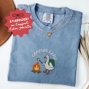 Op de afbeelding: Lichtblauw Comfort Colors T-shirt met een geborduurd ontwerp met een gans die een marshmallow roostert boven een kampvuur. De tekst "CAMPING CREW" staat boven het ontwerp. Het shirt heeft ook de tekst "EMBROIDERY on Comfort Color Shirts" op een rode ster.
